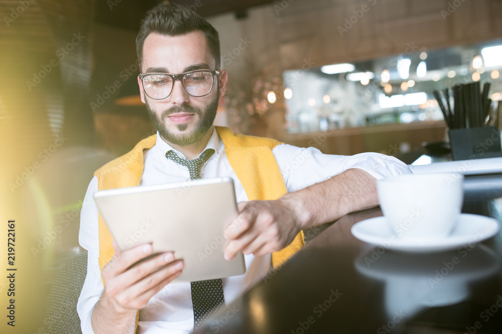 Warm toned portrait of handsome bearded businessman wearing glasses using digital tablet while enjoying coffee break in cafe behind glass wall in sunlight Warm toned portrait of handsome bearded businessman wearing glasses using digital tablet while enjoying coffee break in cafe behind glass wall in sunlight