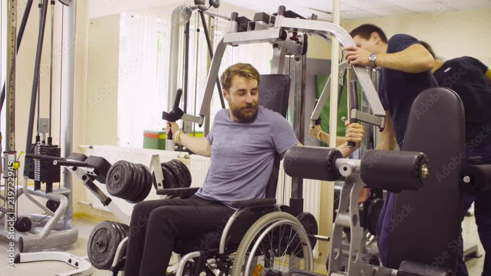 Young disabled man in wheelchair doing strength exercises for hands in the rehabilitation clinic. Doctor adding weight to the simulator. They talking and smiling. Young disabled man in wheelchair doing strength exercises for hands in the rehabilitation clinic. Doctor adding weight to the simulator. They talking and smiling.