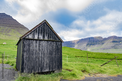 Beautiful view of black faded wooden barn with green grass of sheeps farm and high mountain cliff in the background with cloudy weather sky in Faroe Islands rural