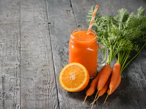 A mug of fresh carrot smoothie with cocktail straw, carrots and oranges on a rustic table.