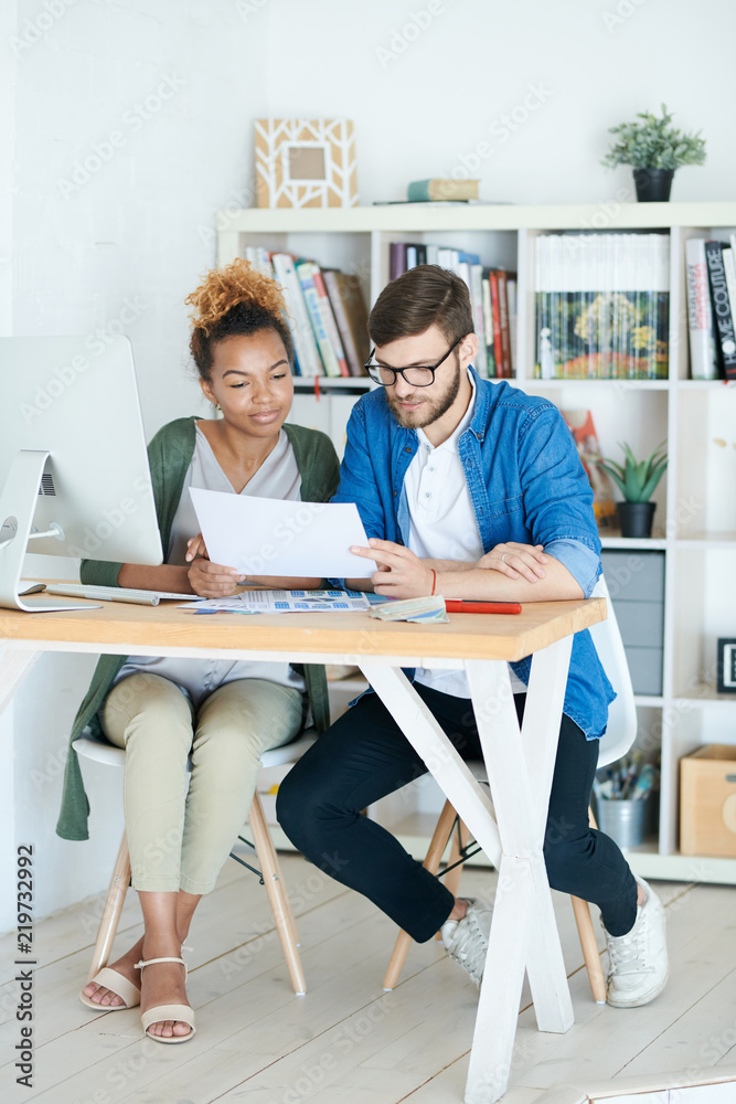 Full length portrait of young African businesswoman discussing documents while working with colleague at desk in modern office, both dressed in casual wear