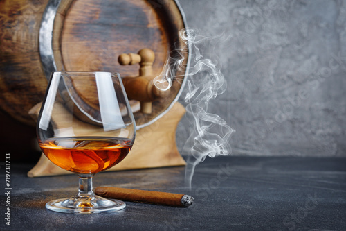 Strong alcoholic drink cognac in sniffer glass with smoking cigar and vintage wooden barrel in cellar