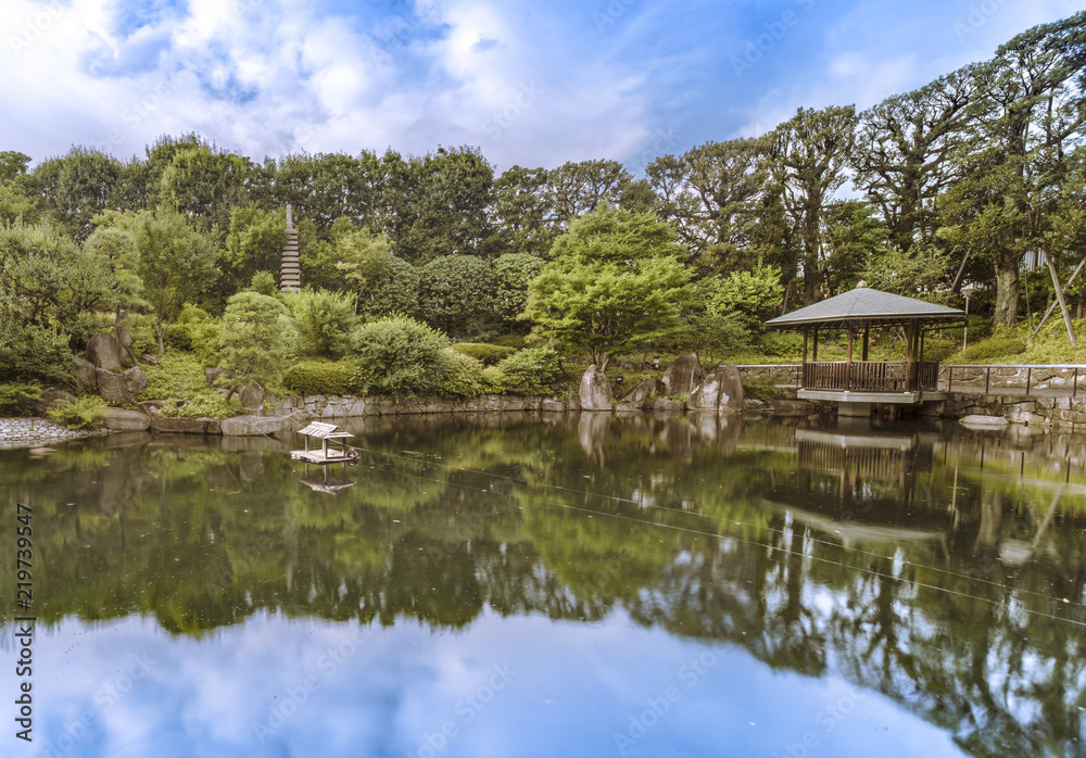 Hexagonal Gazebo in the central pond of Mejiro Garden where ducks are resting and which is surrounded by large rocks and stone pagoda under the foliage of  the pines trees and Momiji maple trees.