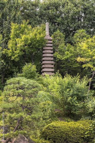 Stone tower pagoda in front of the central pond of Mejiro Garden which is surrounded by large flat stones under the foliage of the Japanese pines trees and a variety of momiji maple trees.