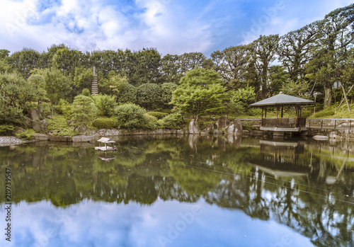 Hexagonal Gazebo in the central pond of Mejiro Garden where ducks are resting and which is surrounded by large rocks and stone pagoda under the foliage of the pines trees and Momiji maple trees.