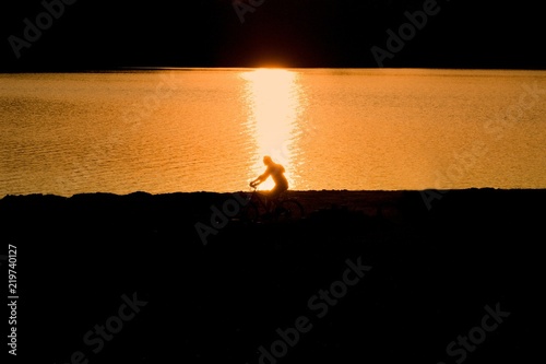 Silhouette of a cyclist with a backpack in sunset and water surface.This is the outline of a man with a backpack on his back on a bicycle in the summer evening at sunset. photo