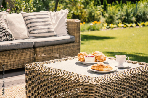 Beige color wicker table and settee on a porch during sunny afternoon in the garden. Croissants and coffee served on the table.