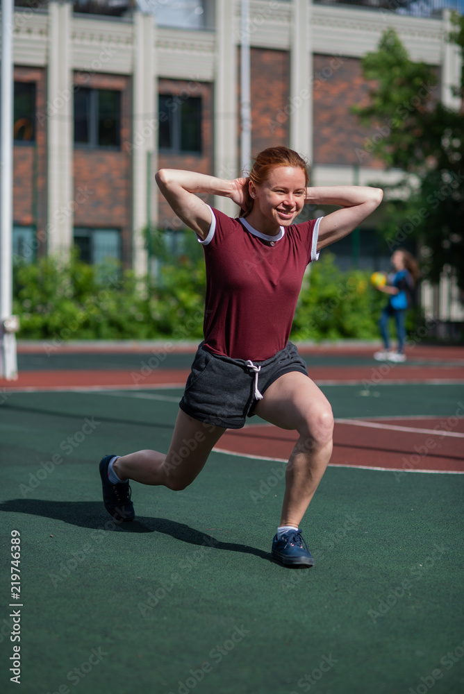 Beautiful young redheaded girl takes a lunge step outdoors. Student engaged in physical education at the stadium