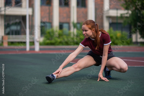 Beautiful young redheaded girl makes a lunge outdoors. Student engaged in stretching in the stadium.
