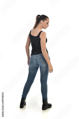 full length portrait of brunette girl wearing black single and jeans. standing pose with back to the camera. isolated on white studio background.
