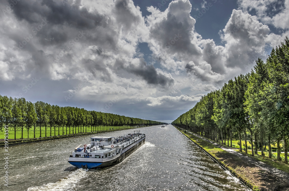 Inland barge on the long straight tree-lined Amsterdam-Rhine canal just south of Amsterdam on a day in summer with dramatic cloud formations - obrazy, fototapety, plakaty 