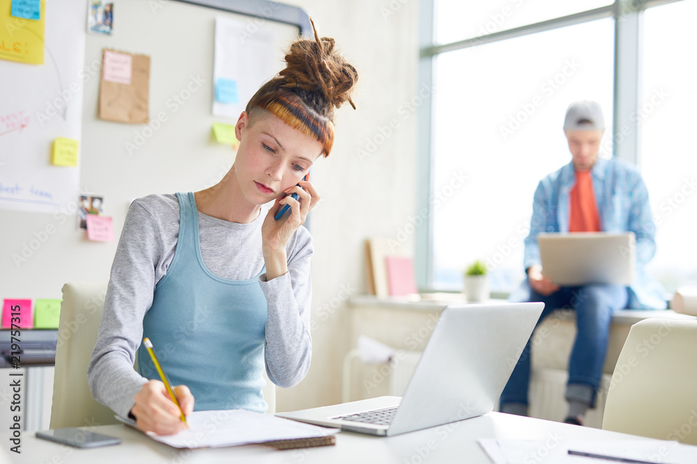 Serious concentrated hipster girl with dreads talking on mobile phone and making notes in sketchpad while working in modern office Serious concentrated hipster girl with dreads talking on mobile phone and making notes in sketchpad while working in modern office