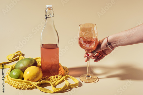 cropped shot of person holding glass with beverage, bottle and string bag with fruits on brown