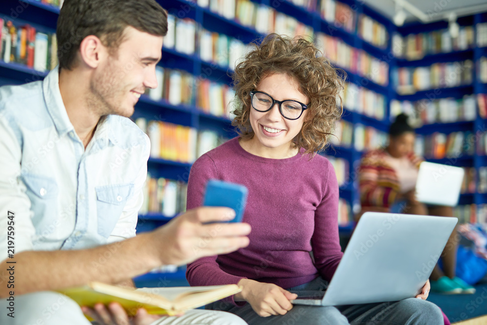 Young man showing something on his smartphone to his classmate while she using laptop in library