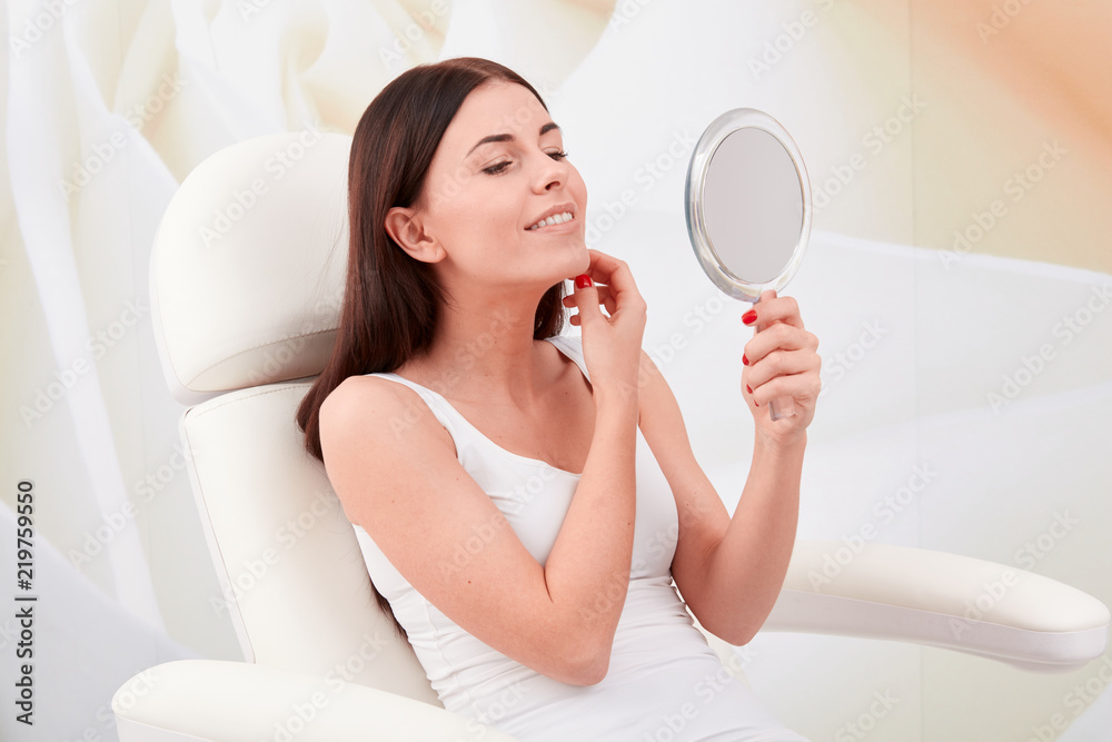 Cosmetic treatments in a professional cosmetic clinic - a young woman looks at her face in the mirror Cosmetic treatments in a professional cosmetic clinic - a young woman looks at her face in the mirror