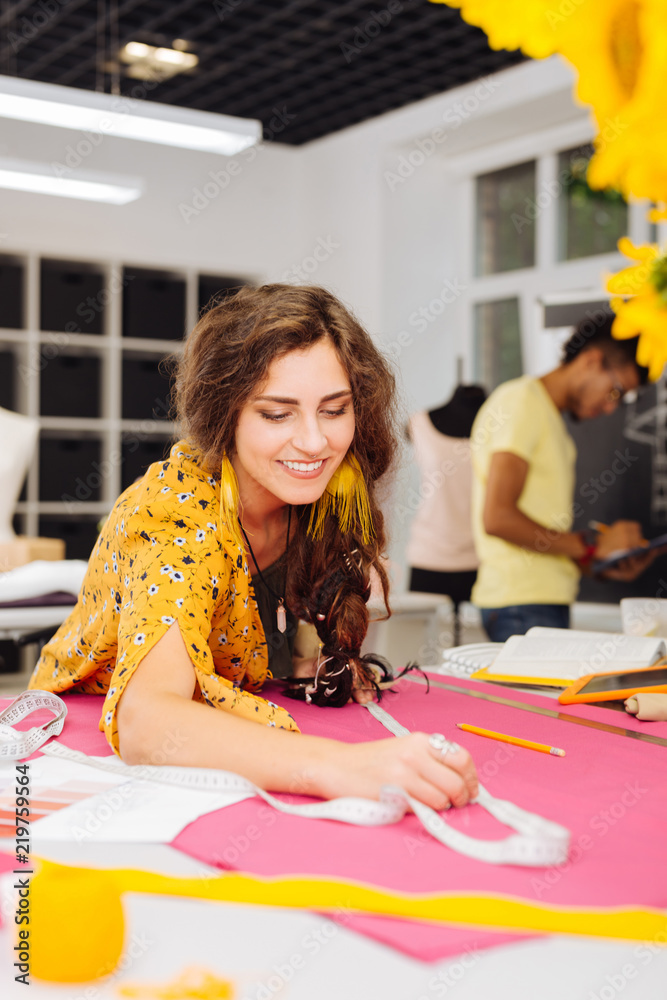 Fototapeta premium Necessary procedure. Cheerful enthusiastic young dressmaker feeling satisfied with her work and smiling while using a tape measure