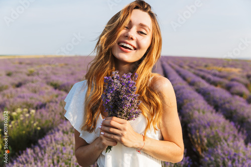 Photo of smiling brunette woman in dress holding bouquet with flowers, while walking outdoor through lavender field in summer