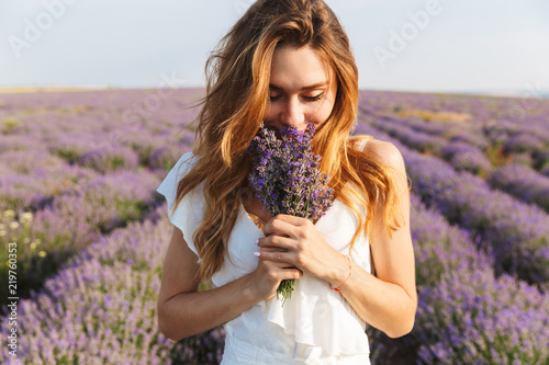 Photo of caucasian young woman in dress holding bouquet of flowers, while walking outdoor through lavender field in summer photo