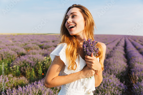 Photo of smiling joyful woman in dress holding bouquet with flowers, while walking outdoor through lavender field in summer