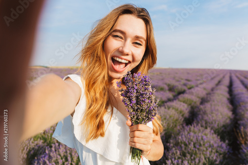 Photo of beautiful young woman in dress taking selfie while holding bouquet with flowers, during walk through lavender field in summer