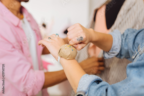 Taking pins. Careful young dressmaker standing next to her colleague and taking colorful pins from the pin cushion on her wrist