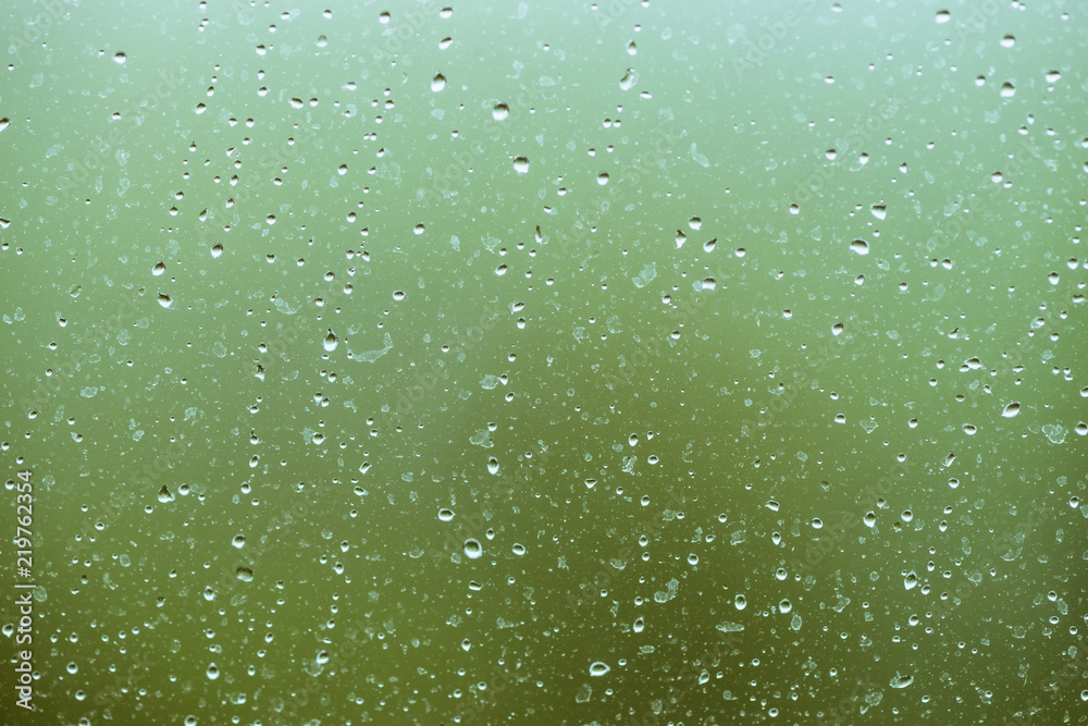 Dirty glass with drops of rain. Raindrops on green clear bokeh background. Greenery outside window. Droplets and stains close up. Detailed transparent texture in macro.