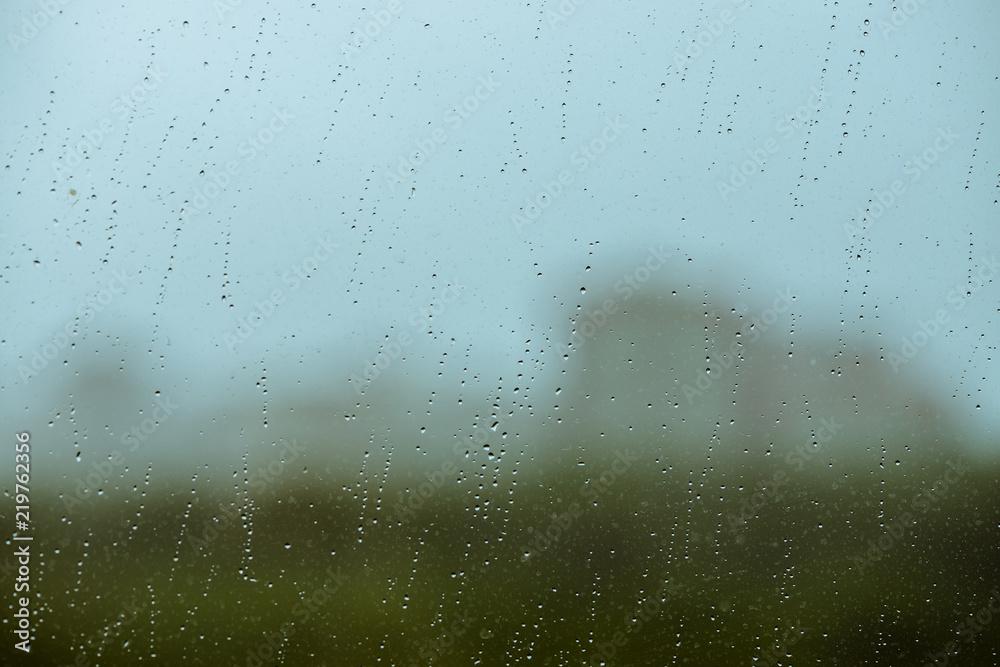 Dirty glass with drops of rain. Raindrops on background from greenery, building and sky in bokeh. City outside window. Droplets and stains close up. Detailed transparent texture in macro.