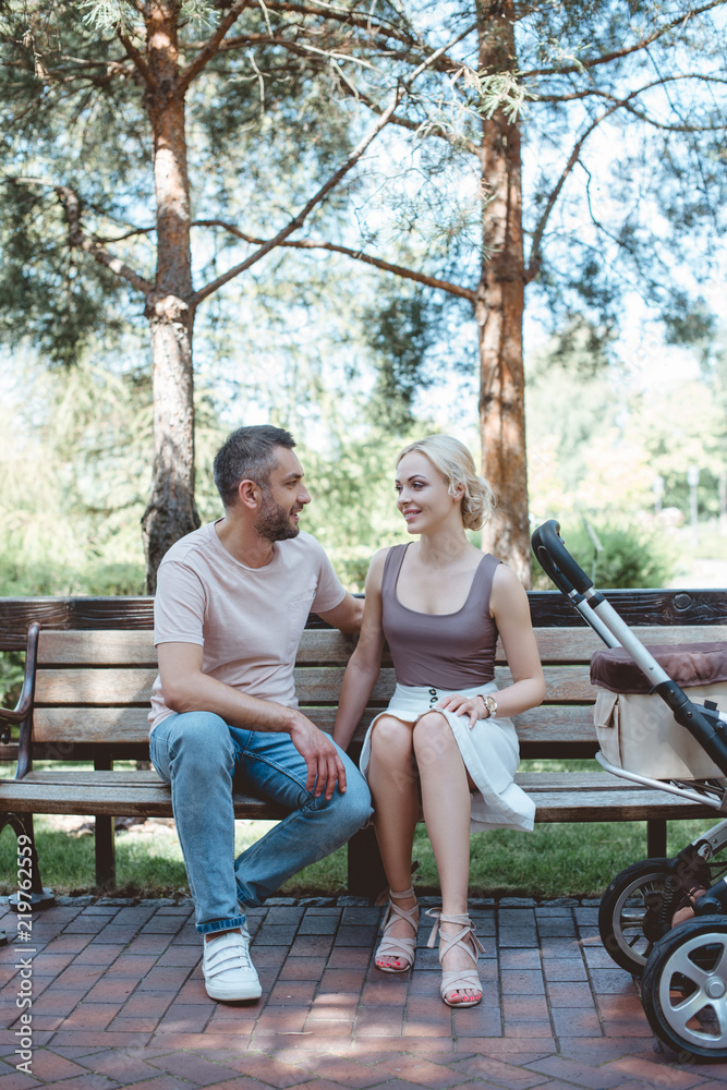 parents sitting on bench near baby carriage in park and looking at each other parents sitting on bench near baby carriage in park and looking at each other