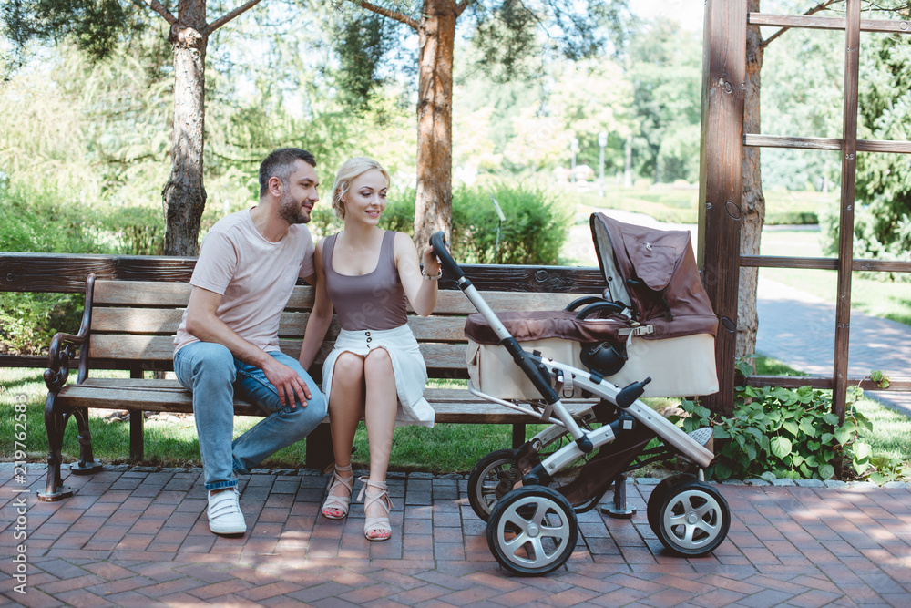father and mother sitting on bench near baby carriage in park