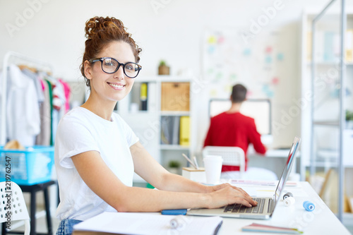 Happy young manager or designer sitting by desk in front of laptop, typing and looking at camera with colleague on background