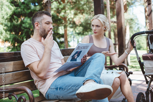 husband smoking cigarette and reading newspaper near baby carriage in park, angry wife looking at him