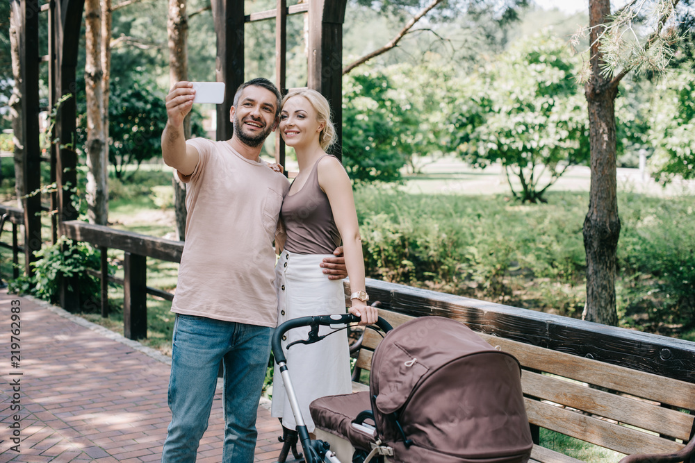 mother and father taking selfie with smartphone near baby carriage in park mother and father taking selfie with smartphone near baby carriage in park