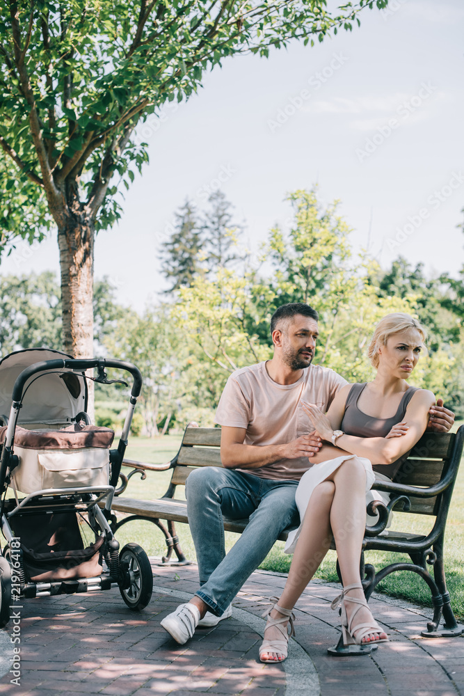 husband hugging irritated wife on bench near baby carriage in park husband hugging irritated wife on bench near baby carriage in park
