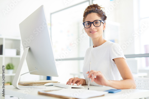 Young confident manager or designer sitting by her workplace in office in front of computer monitor