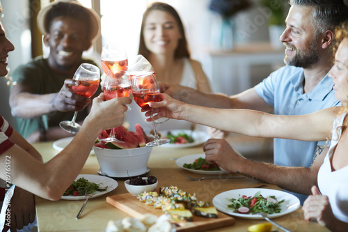 Group of young cheerful friends clinking with red drinks in wineglasses over served table at party