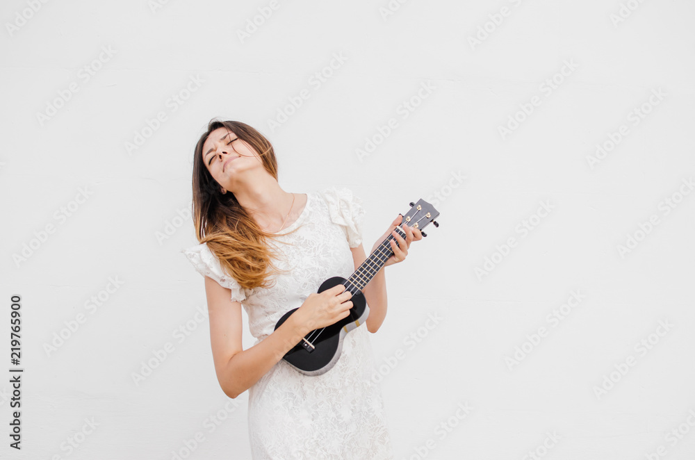 Happy, cute young woman plays ukulele against the background of a white wall and smiles. Happy, cute young woman plays ukulele against the background of a white wall and smiles.