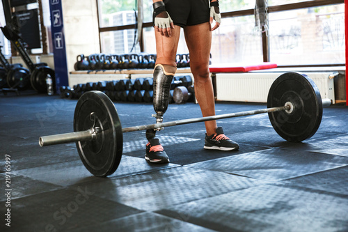 Cropped image of athletic caucasian disabled woman wearing prosthesis in tracksuit, training and lifting barbell in gym