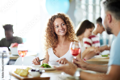Smiling young woman eating salad and talking to man by her table while enjoying party with friends