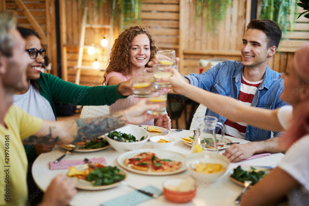 Young excited friends clinking with drinks over served festive table while celebrating life event