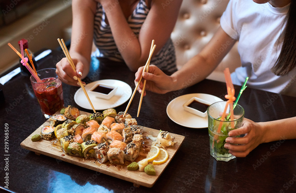 Two yang girls are eating delicious rolling sushi at restaurant served on the wooden board with chopsticks, soy sauce and cocktails