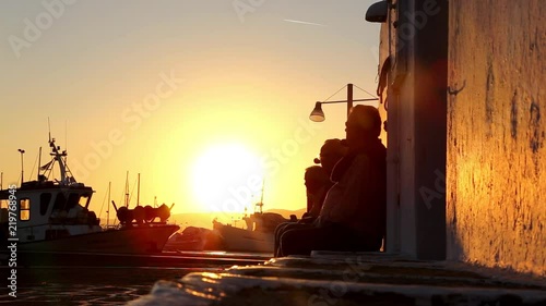 Silhouette of group of unrecognizable senior people talking and relaxing on a bench by the port ovelooking the fishing baots in Chania, Crete island, Greece at sunset. photo