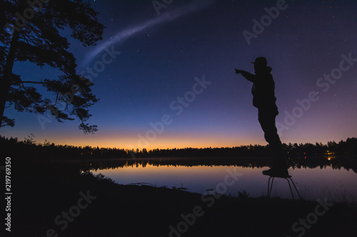 A man silhouette at the still lake in night time with a late sunset in background. Different poses towards dark blue sky.  © Viesturs