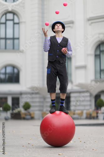 The clown and balancer juggles with pink balls , standing on a big red ball in the street of a European city