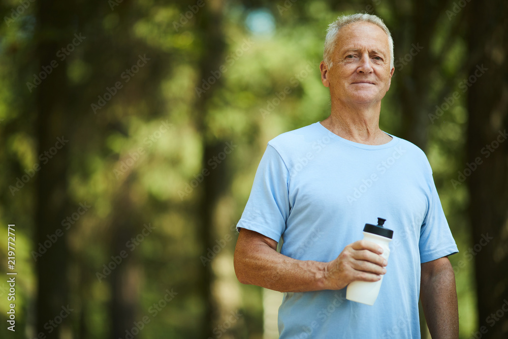 Sporty senior man with bottle of water standing in park or forest on summer day and having refreshment Sporty senior man with bottle of water standing in park or forest on summer day and having refreshment