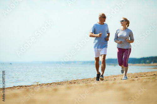 Active senior man and woman running down sandy beach with waterside on background