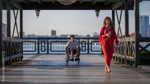 A beautiful girl with red hair in a red dress walks along the waterfront and behind her a young guy rides in a wheelchair