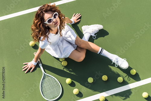 overhead view of fashionable tennis player in white sportswear and sunglasses resting on tennis court with balls and racket near by © LIGHTFIELD STUDIOS