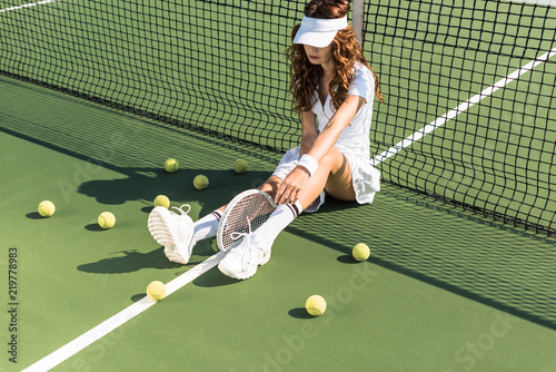 beautiful tennis player in white tennis uniform with racket sitting near tennis net on court with tennis balls around © LIGHTFIELD STUDIOS