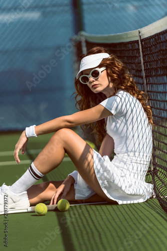 side view of fashionable female tennis player in sunglasses resting near net on tennis court with equipment near by
