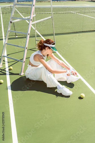 side view of young woman in stylish white clothing and cap tying shoelaces on tennis court with racket and balls © LIGHTFIELD STUDIOS